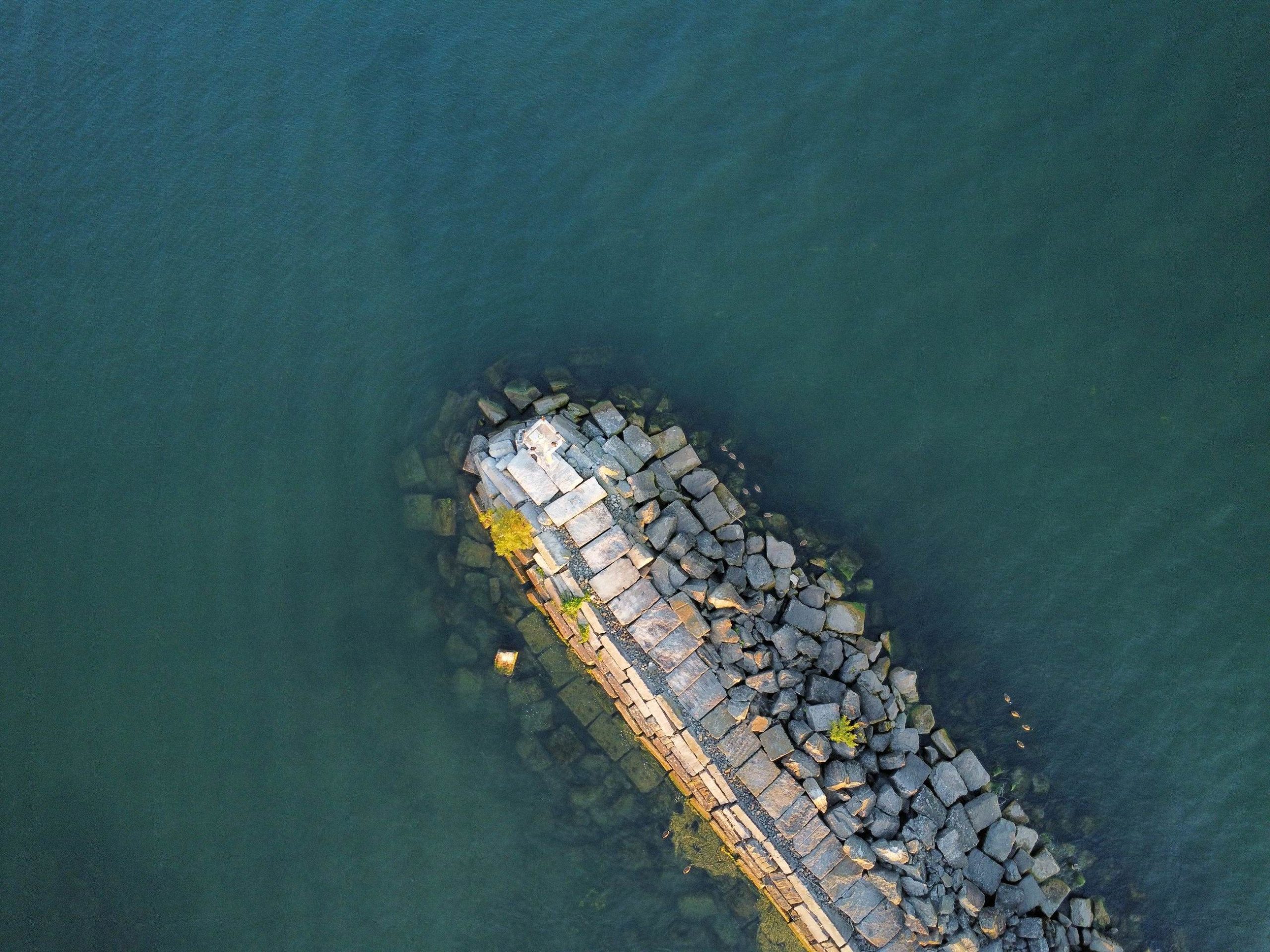 A rock breakwater from above, reaching into deep blue-green water